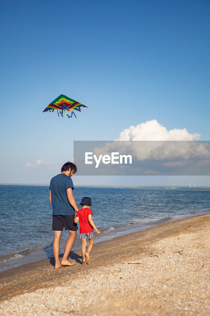 beach, sky, land, water, sea, two people, nature, men, holiday, togetherness, childhood, vacation, trip, full length, sand, child, adult, leisure activity, family, horizon over water, ocean, kite - toy, horizon, summer, body of water, emotion, women, blue, coast, parent, day, casual clothing, bonding, shore, positive emotion, happiness, cloud, one parent, fun, female, copy space, motion, carefree, enjoyment, father, love, standing, beauty in nature, toy, relaxation, outdoors, lifestyles, travel, sunlight, sunny, travel destinations, person, kite sports, smiling, holding, clothing, scenics - nature, barefoot, looking