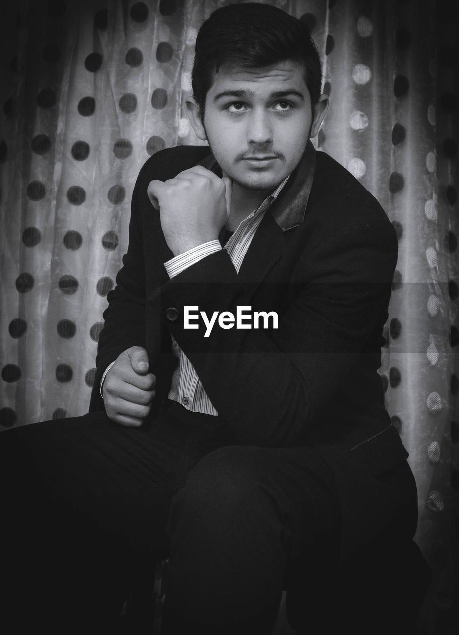 black, black and white, one person, portrait, adult, monochrome photography, monochrome, young adult, sitting, looking at camera, men, indoors, darkness, white, formal wear, person, lifestyles, photo shoot, business, serious, clothing, three quarter length, front view, portrait photography, looking, fashion