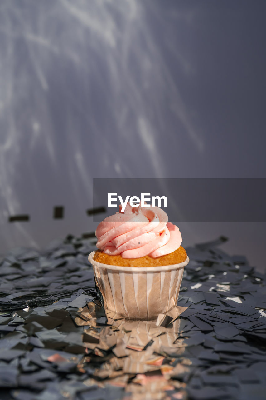A cupcake with a pink cap stands among the flying silver confetti.