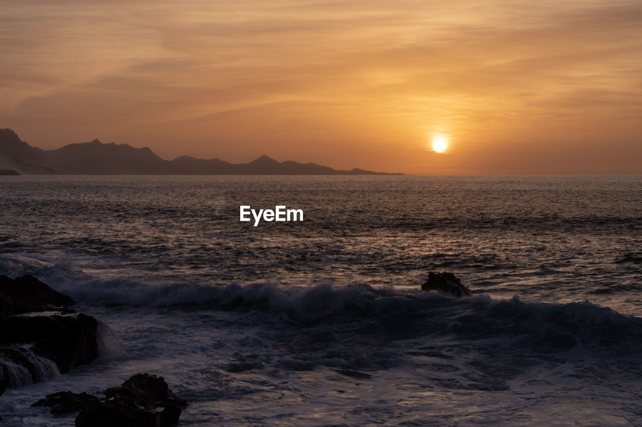sky, sea, water, sunset, beauty in nature, scenics - nature, land, ocean, nature, wave, beach, shore, coast, horizon, sun, cloud, motion, wind wave, tranquility, rock, dawn, environment, body of water, tranquil scene, sunlight, dramatic sky, orange color, sports, seascape, idyllic, horizon over water, landscape, outdoors, travel destinations, evening, no people, silhouette, travel, sand, non-urban scene, water sports, tourism