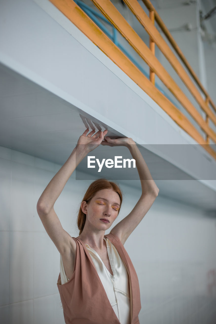 Teenage girl with arms raised and eyes closed standing against wall