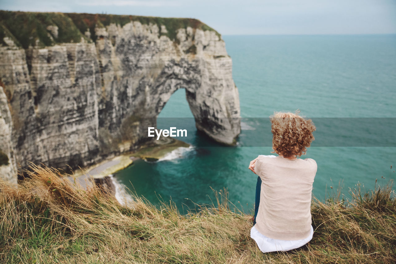 Rear view of woman sitting on field by sea against sky