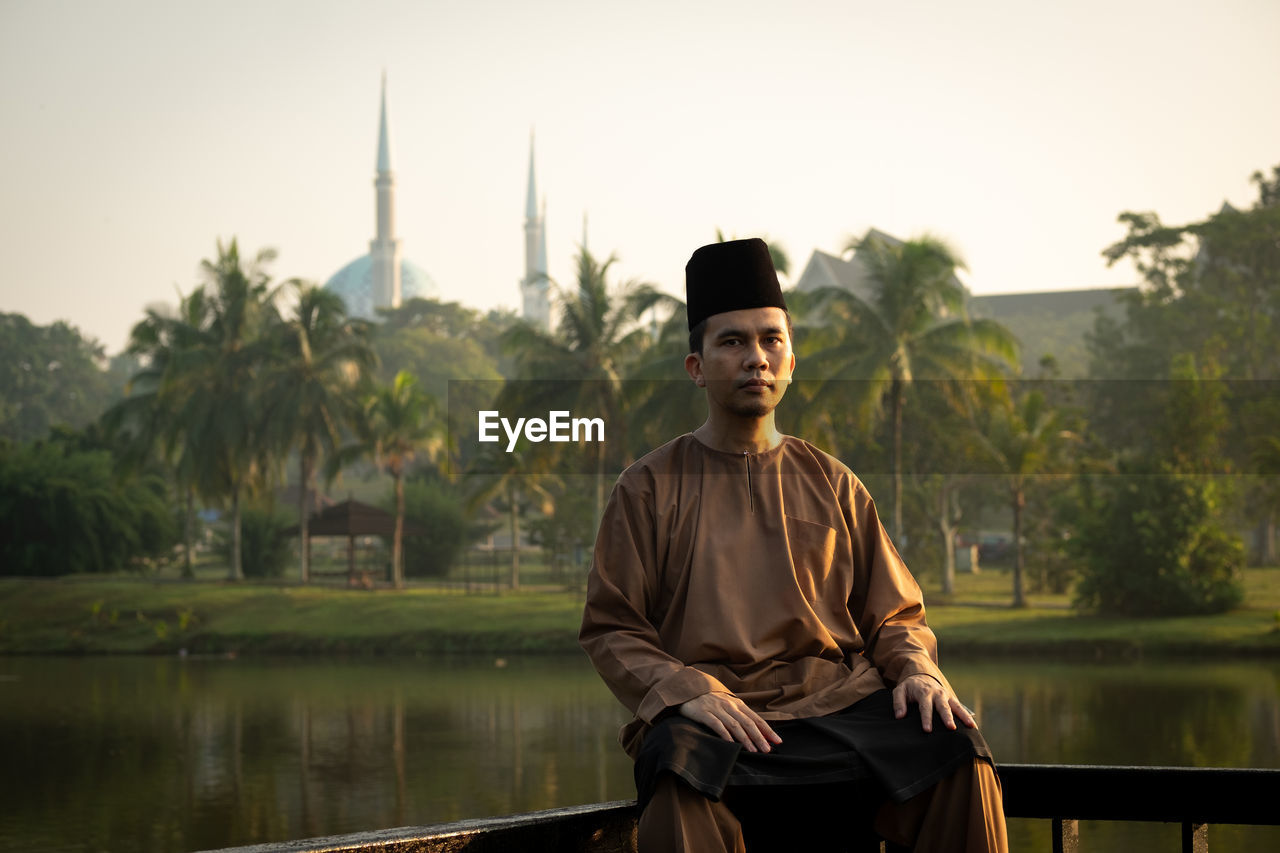 Portrait of man wearing traditional clothing while sitting on railing against lake during sunset