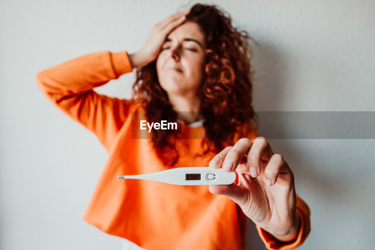 Sick mid adult woman showing thermometer while standing against wall