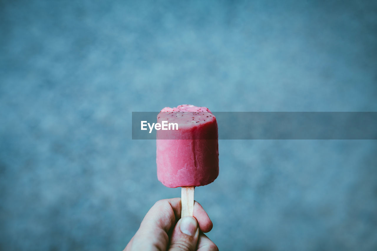 Crop person showing tasty berry ice pop of pink color on blurred blue background person