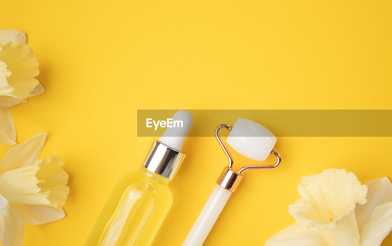Unbranded serum bottle, face roller and narcissus flowers on yellow background. cosmetic facial skin 