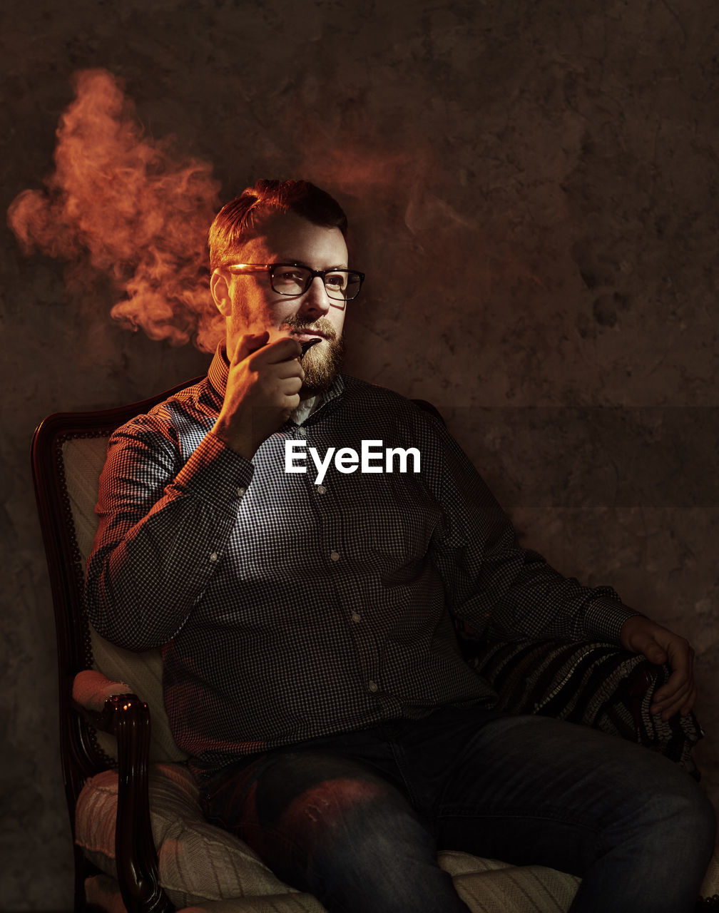 Mature man looking away while smoking on chair in darkroom at home