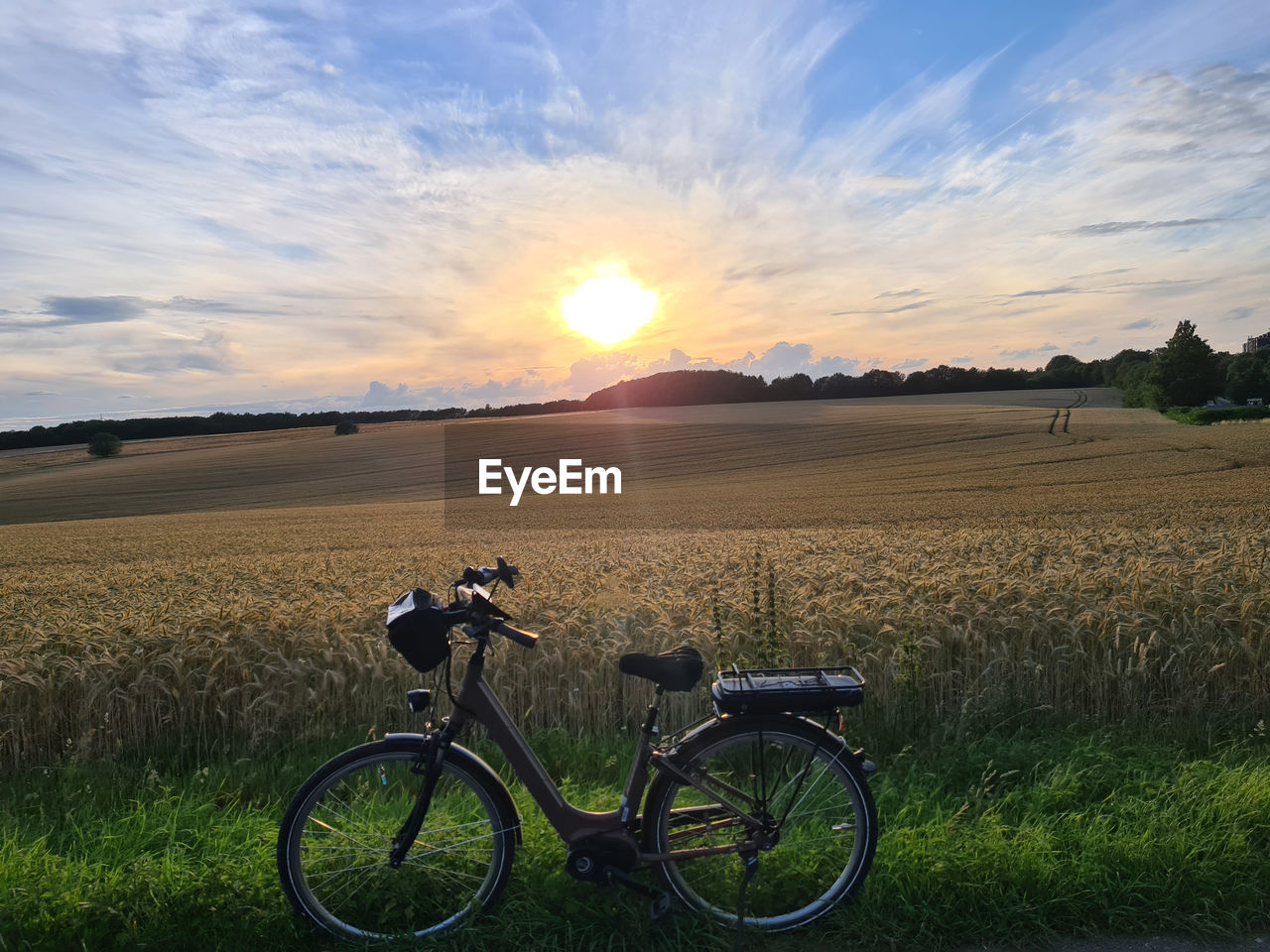 sky, landscape, transportation, field, sunset, environment, nature, land, bicycle, rural scene, plant, cloud, sunlight, beauty in nature, scenics - nature, agriculture, grass, mode of transportation, rural area, crop, horizon, sun, land vehicle, tranquility, lens flare, tranquil scene, vehicle, sunbeam, cereal plant, non-urban scene, travel, outdoors, farm, prairie, summer, horizon over land, road, idyllic, no people, dusk, back lit, sports equipment, plain, bicycle wheel, growth, grassland, dramatic sky, day, activity