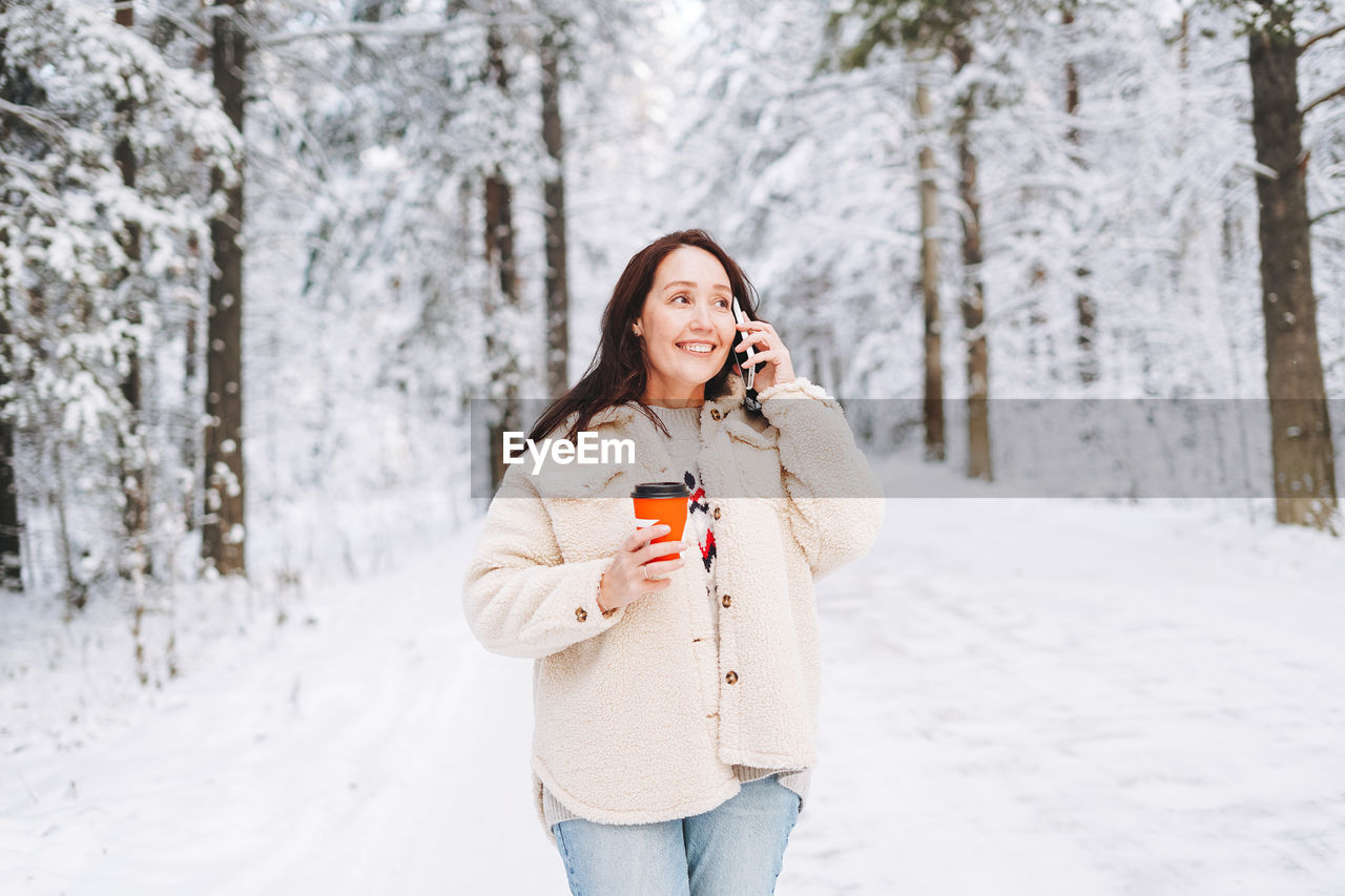 Smiling woman with dark hair in winter clothes using mobile phone in snowy winter forest