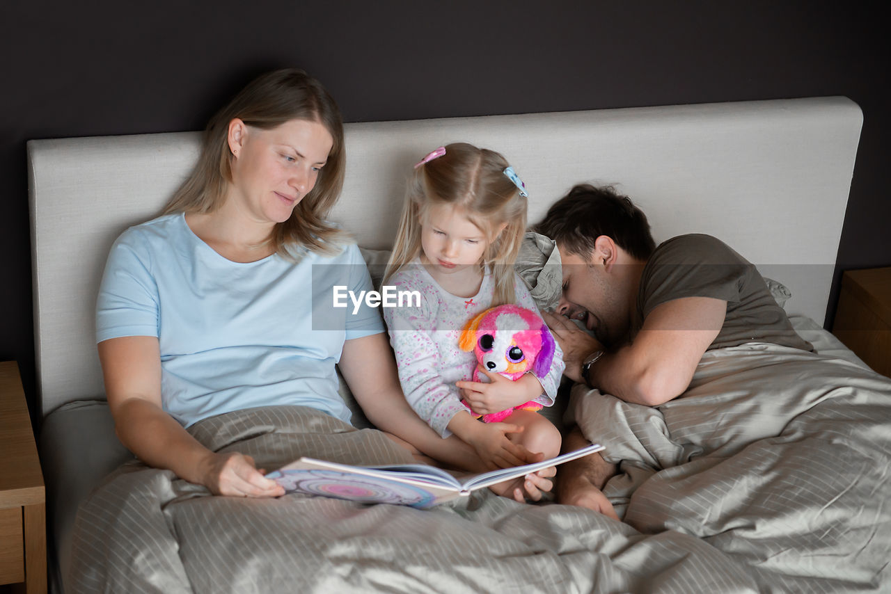 High angle view of mother reading book for daughter while father sleeping on bed at home