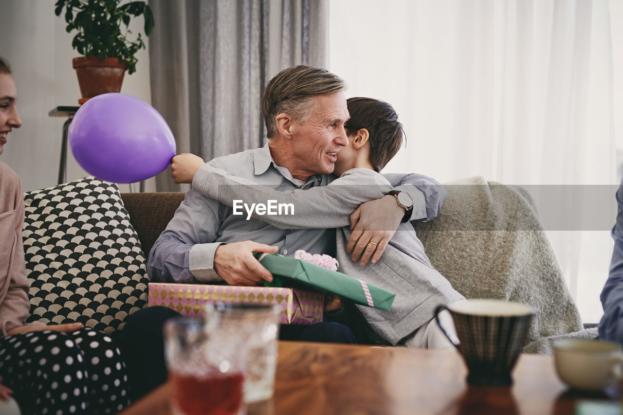 Grandfather holding birthday gift while embracing grandson on sofa at home