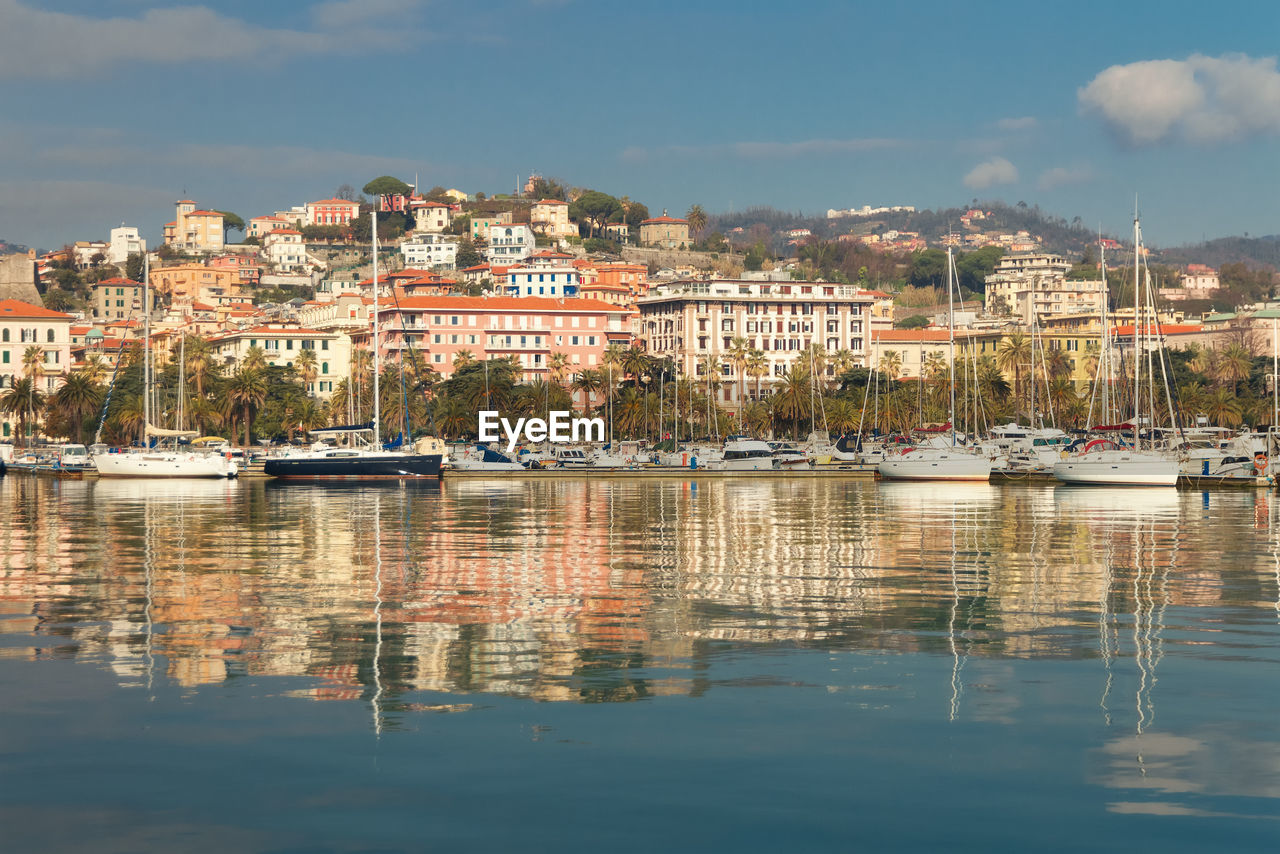 SAILBOATS MOORED IN HARBOR BY BUILDINGS AGAINST SKY