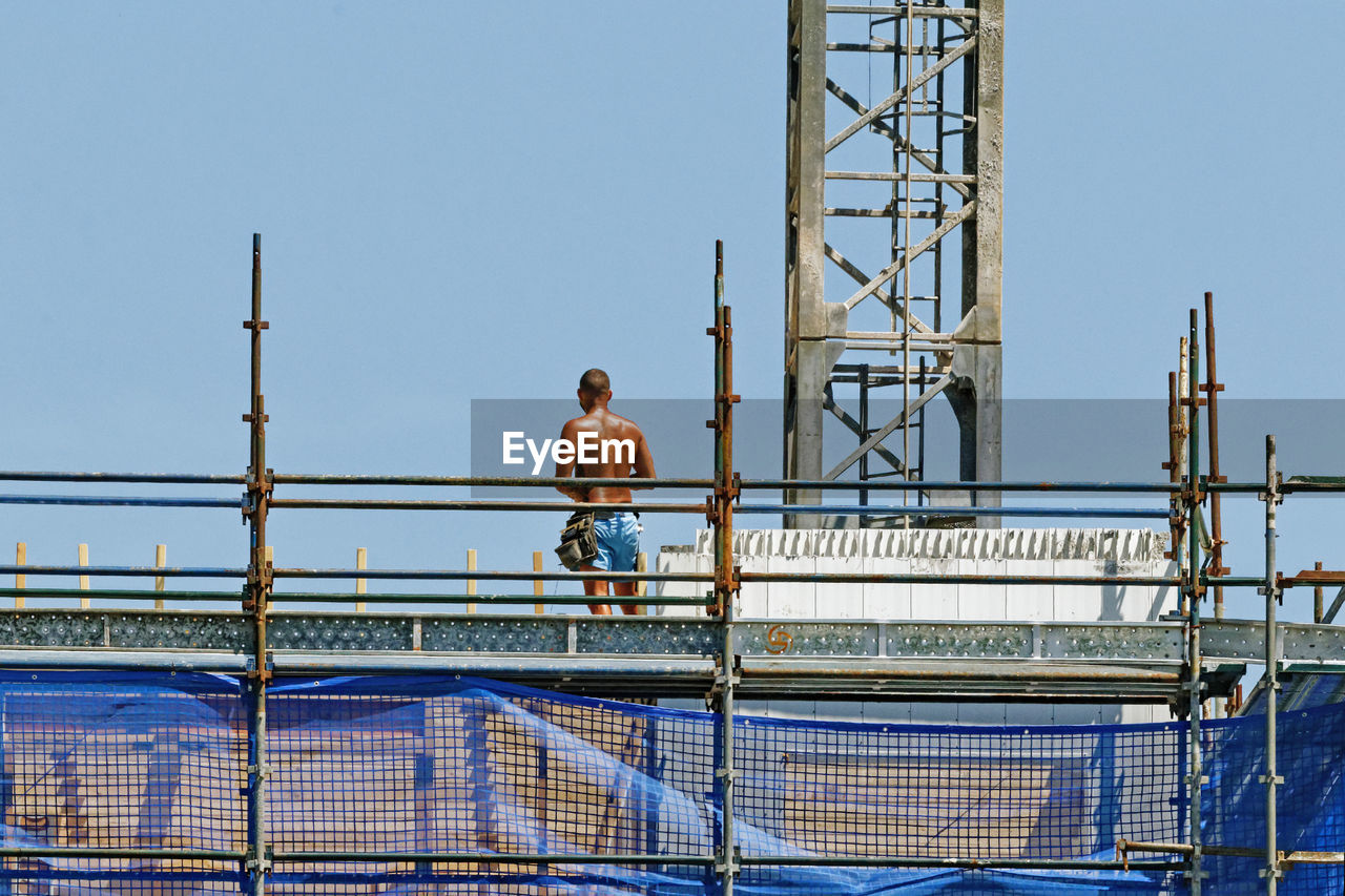 REAR VIEW OF SHIRTLESS MAN STANDING AGAINST RAILING