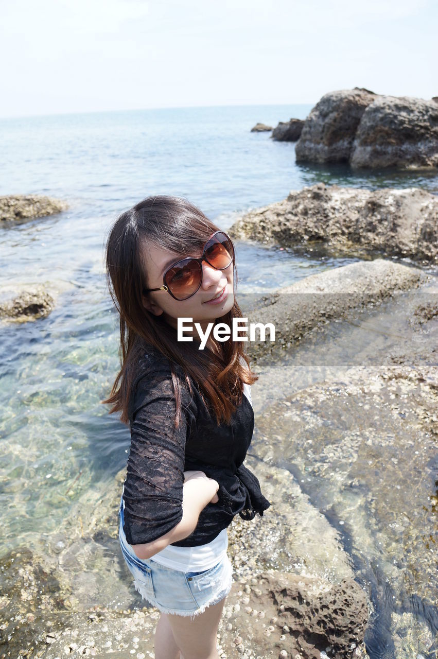 Portrait of smiling young woman standing on rock at beach against sky