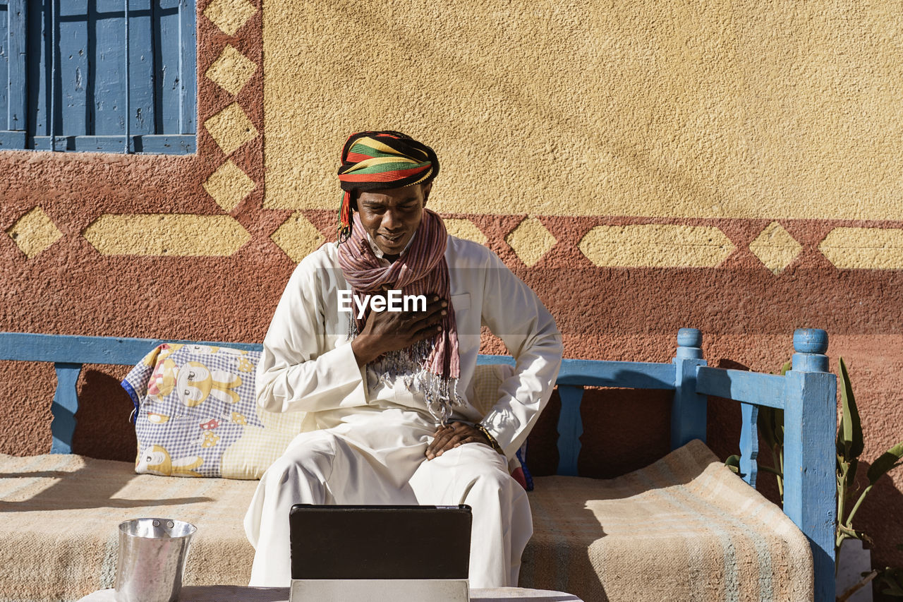 Sincere egyptian male in traditional turban and robe having video chat on tablet while sitting on sofa in backyard on sunny day