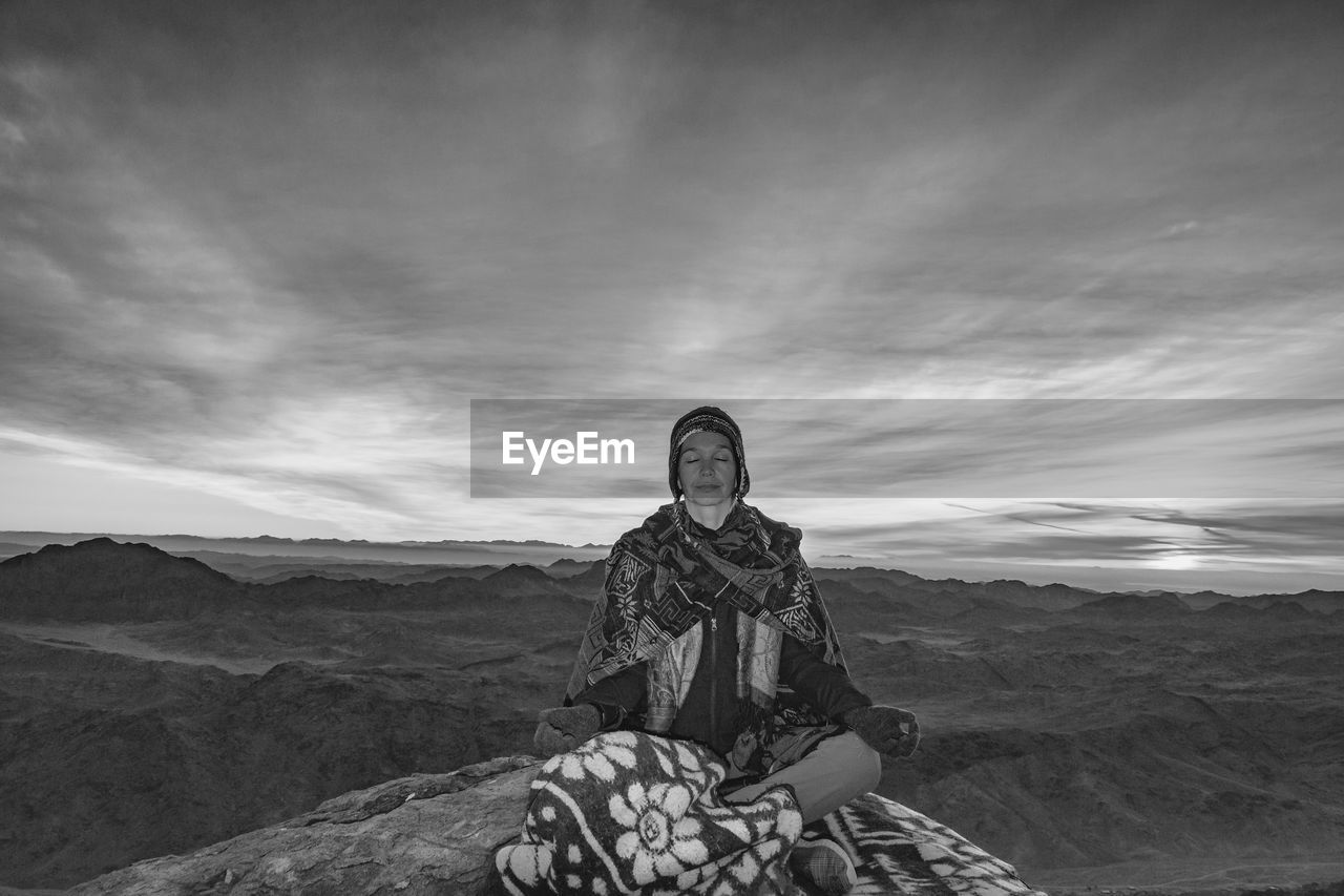 Woman meditating while sitting on rock against sky during sunset