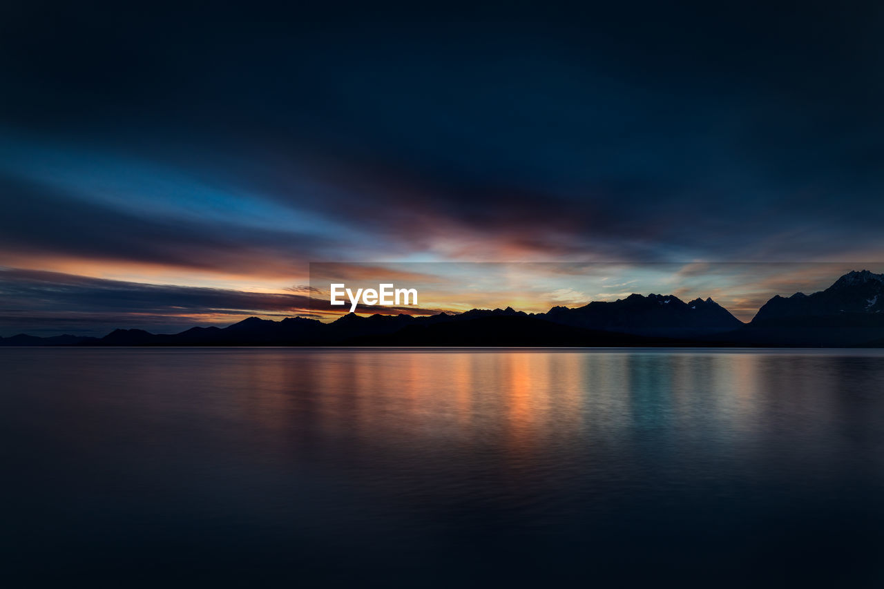 SCENIC VIEW OF LAKE BY SILHOUETTE MOUNTAINS AGAINST SKY AT SUNSET
