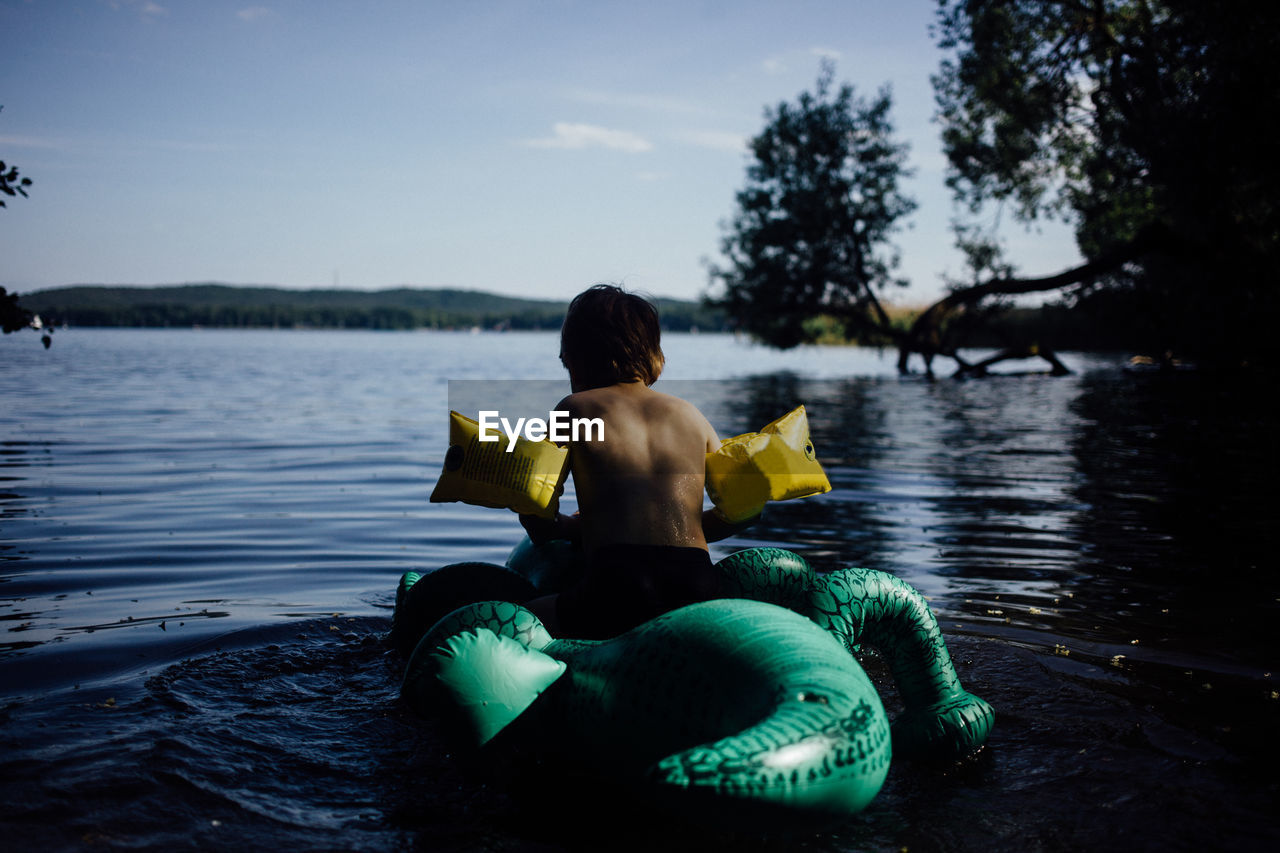 Rear view of boy sitting on inflatable crocodile at lake against sky