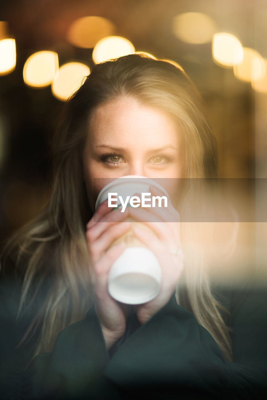 Portrait of young woman drinking coffee at cafe seen through glass