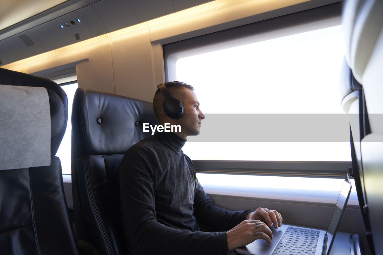 Side view of young male freelancer sitting on passenger seat in modern train and typing on netbook while working during trip