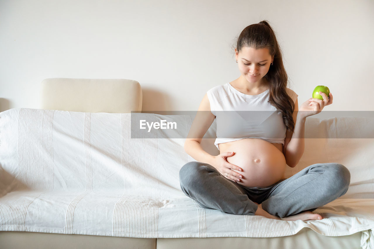 Young pregnant woman holding fruit while sitting on sofa at home