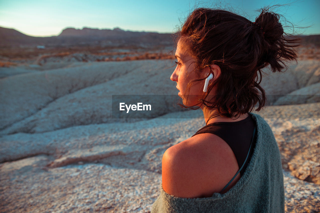 Side view of young female in sportive outfit and listening to music with wireless earbuds contemplating amazing scenery of rough stony badlands at sunset time