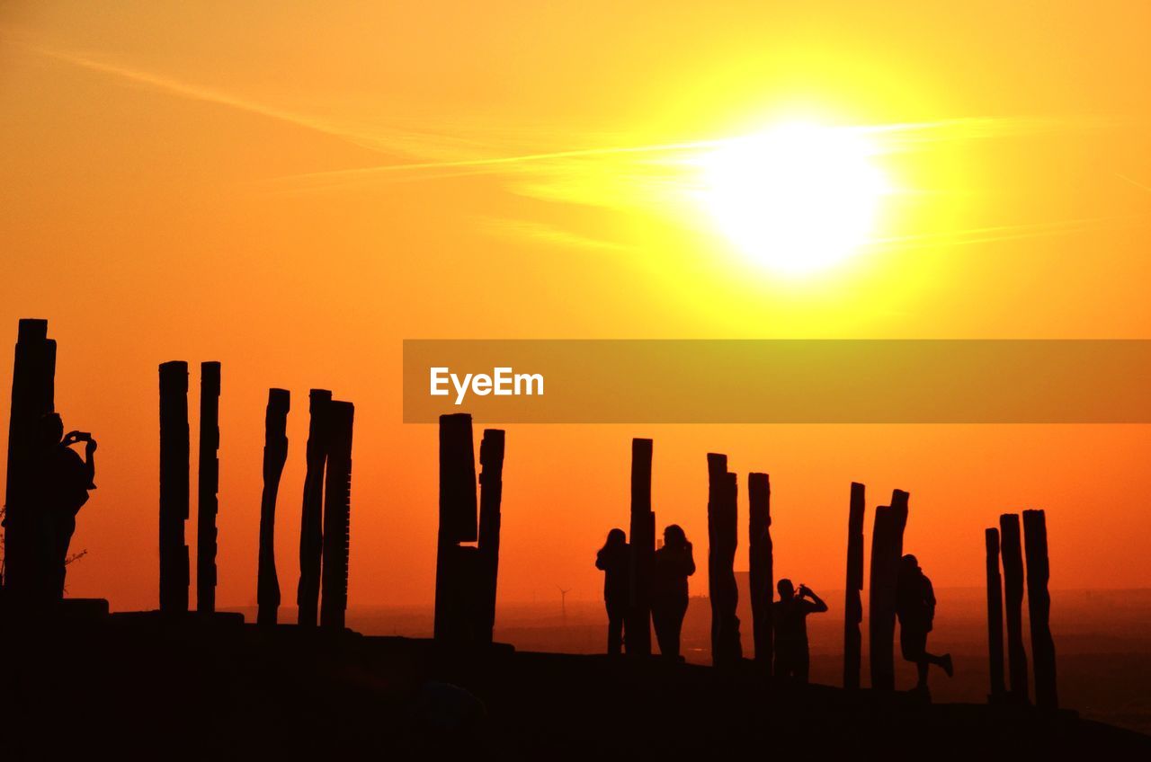 Silhouette people standing amidst wooden posts against sky during sunrise
