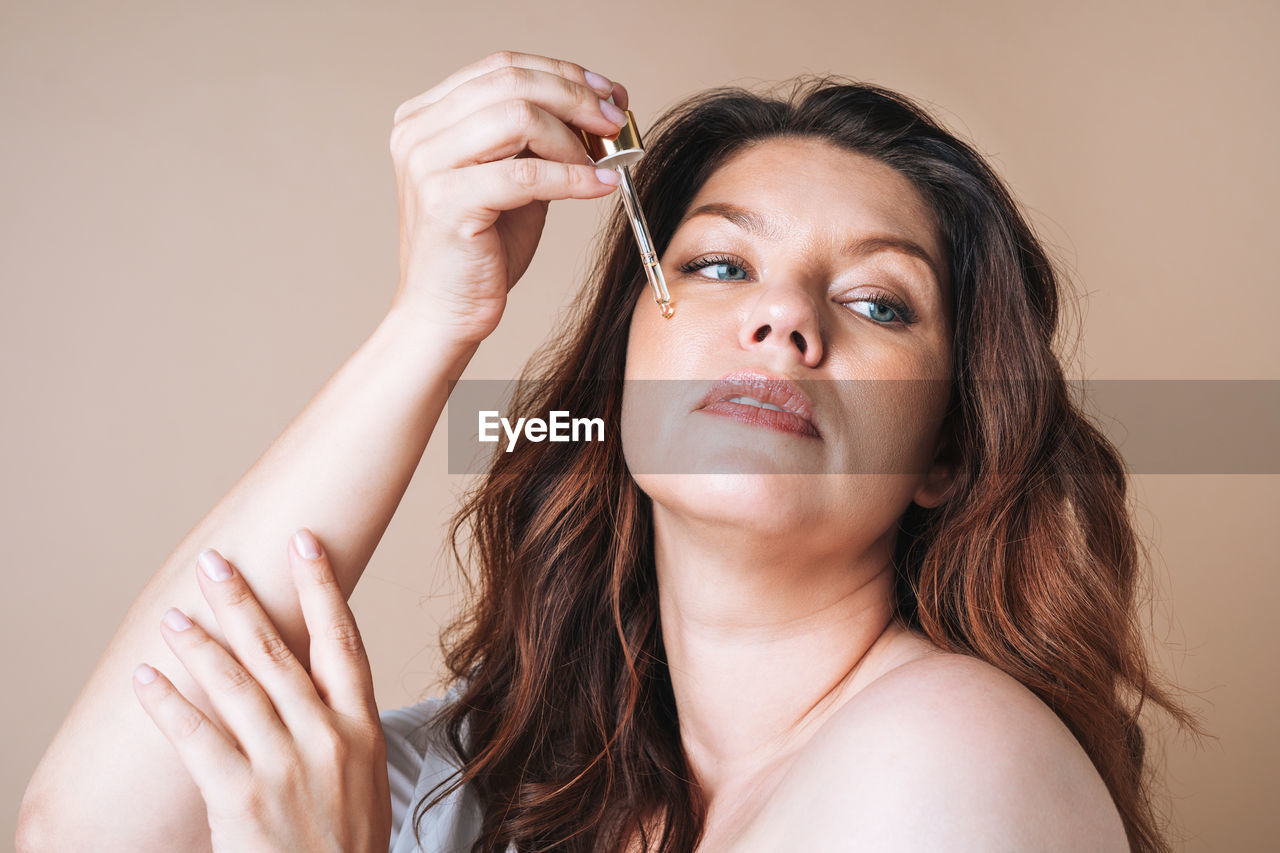 Beauty portrait of body positive woman with dark hair with pipette with oil on beige background