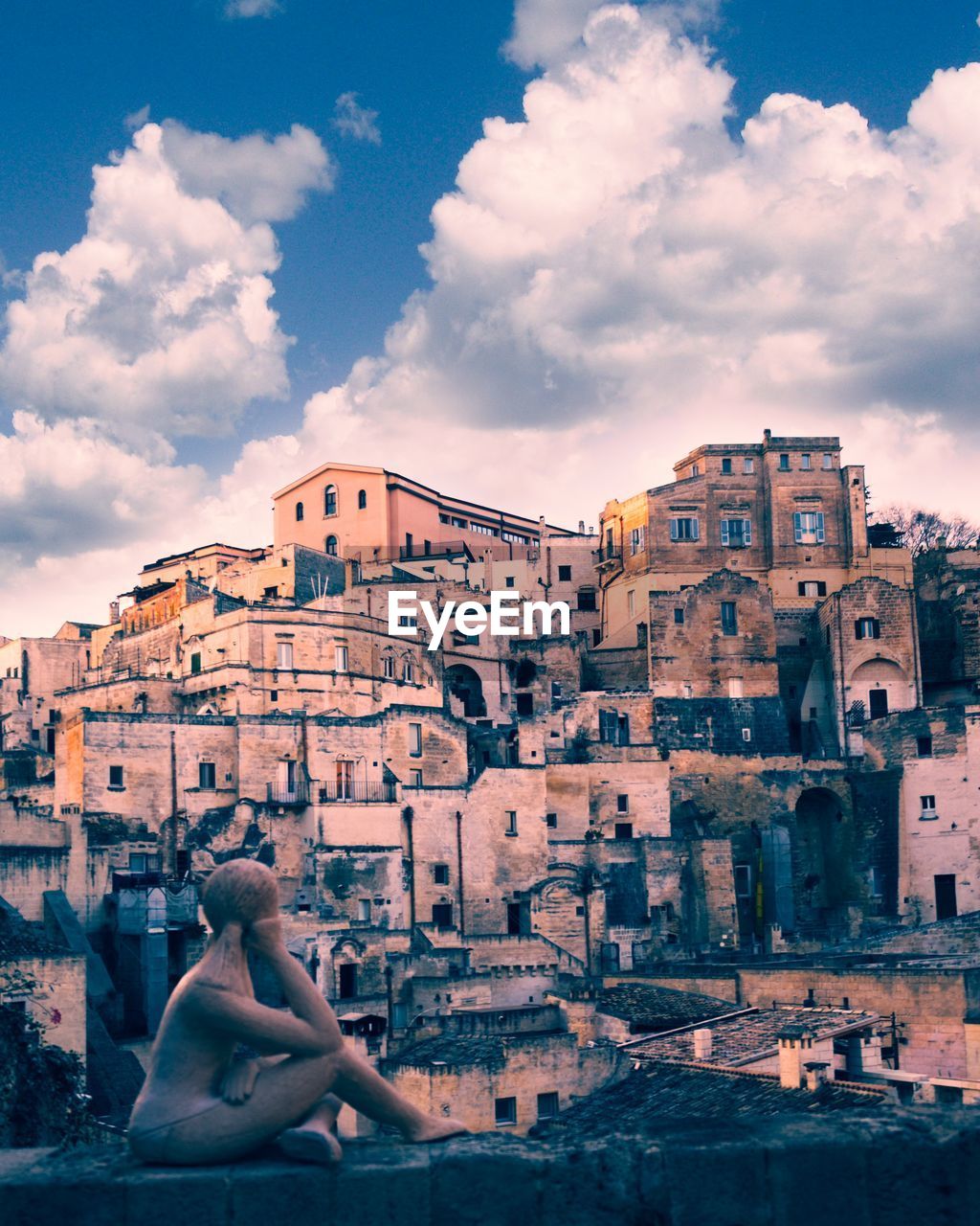 WOMAN SITTING BY BUILDINGS AGAINST SKY