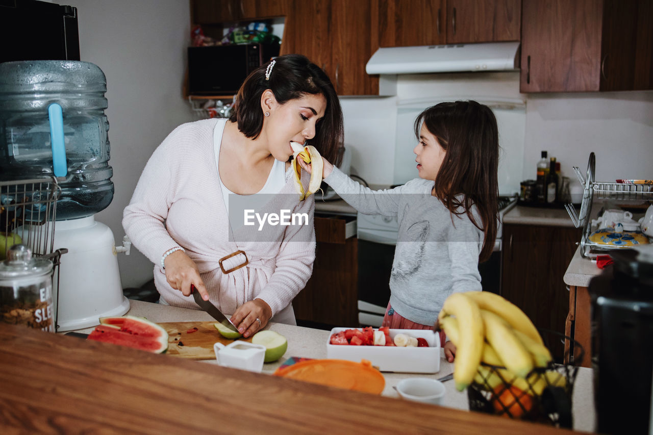 Cute little girl feeding ripe banana to mother cutting fresh fruits on at table with various food while standing in kitchen at home