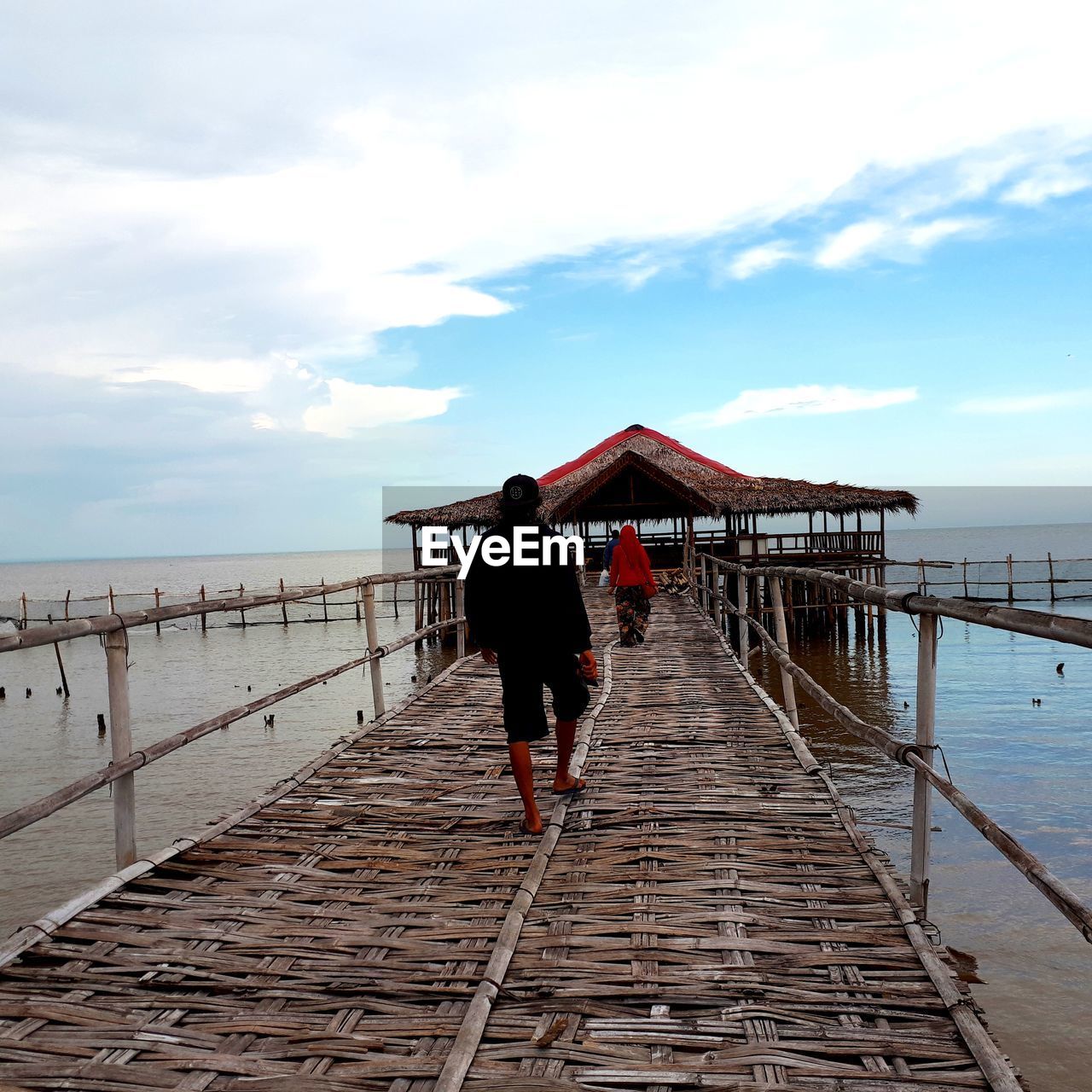 Rear view of man and woman walking on pier over sea against cloudy sky