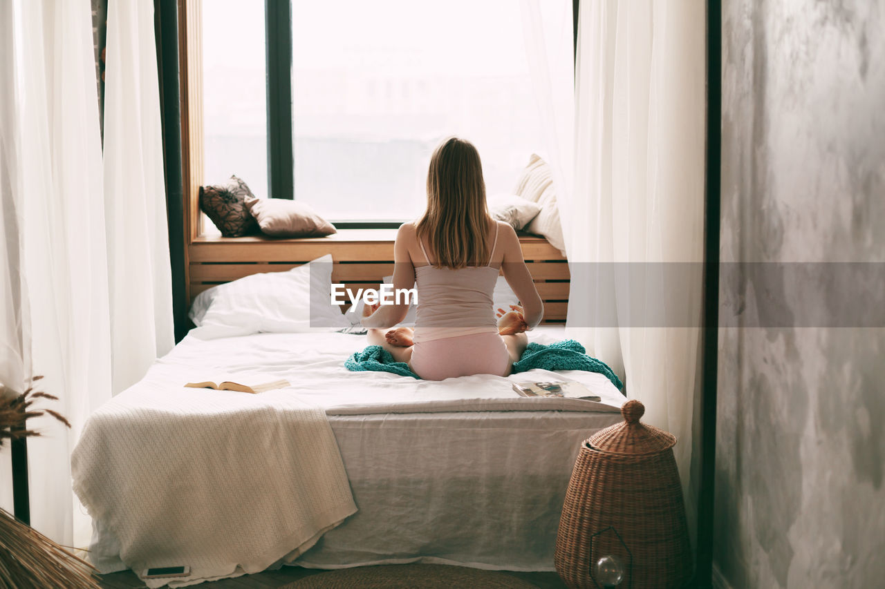 A young woman in pyjamas is sitting on a bed in a lotus position 