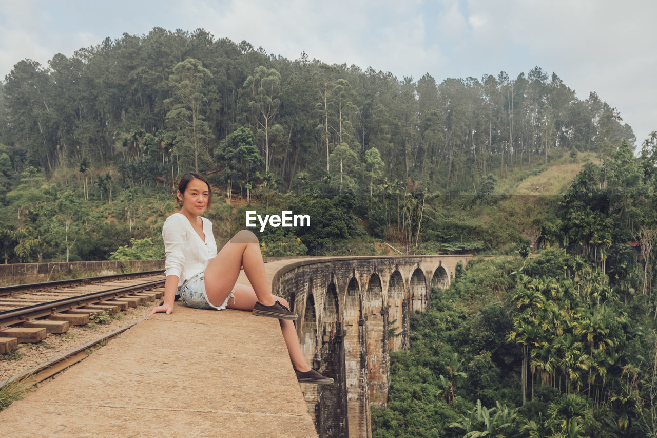 Side view of asian lady in casual wear sitting on edge of old bridge and looking at camera with green forest on background in sri lanka asia