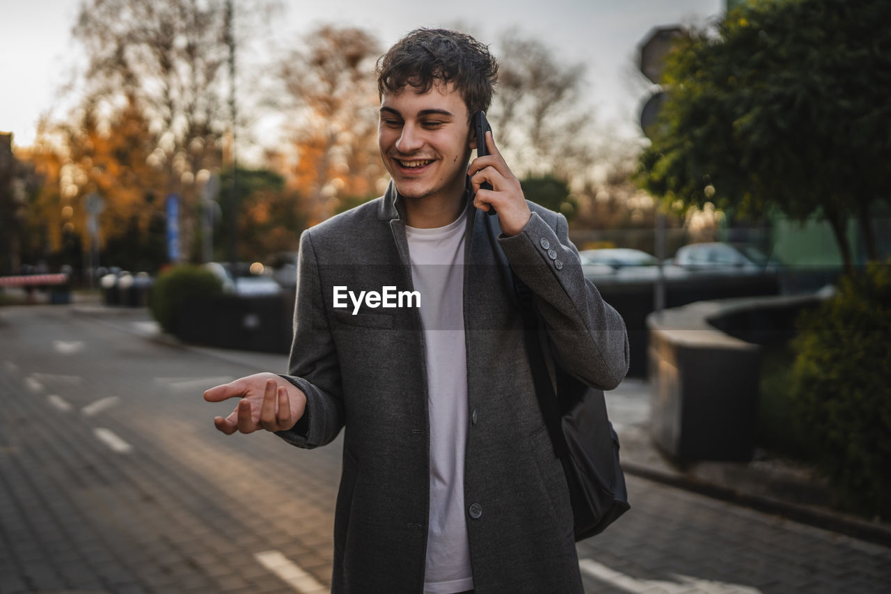 one person, city, adult, smiling, men, young adult, happiness, street, architecture, wireless technology, businessman, communication, technology, emotion, mobile phone, city life, standing, cheerful, business, telephone, lifestyles, formal wear, smartphone, portable information device, front view, clothing, looking, transportation, smile, using phone, nature, waist up, spring, teeth, outdoors, listening, day, blazer, city street, enjoyment, business finance and industry, motion, focus on foreground, on the move, portrait, road, tree, leisure activity, copy space, portability, footpath, beard, outerwear, corporate business, coat, headphones