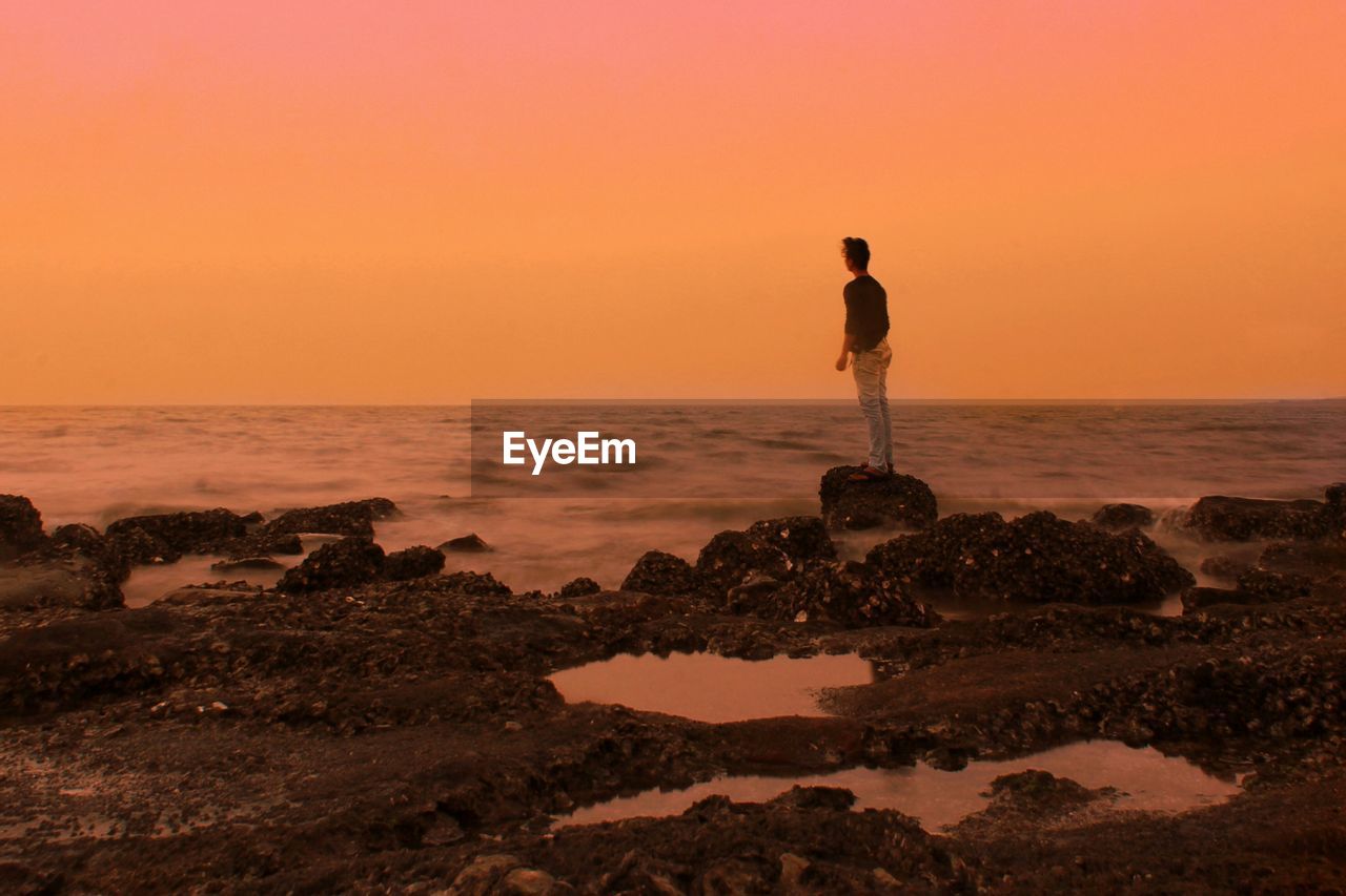 Rear view of man standing on rock at seashore against sky during sunset