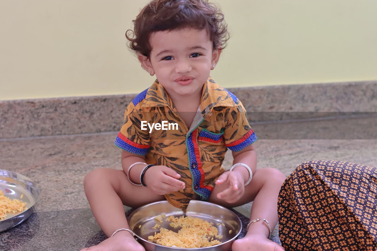 A child sits inside the house eating food and the little boy has a plate full of food