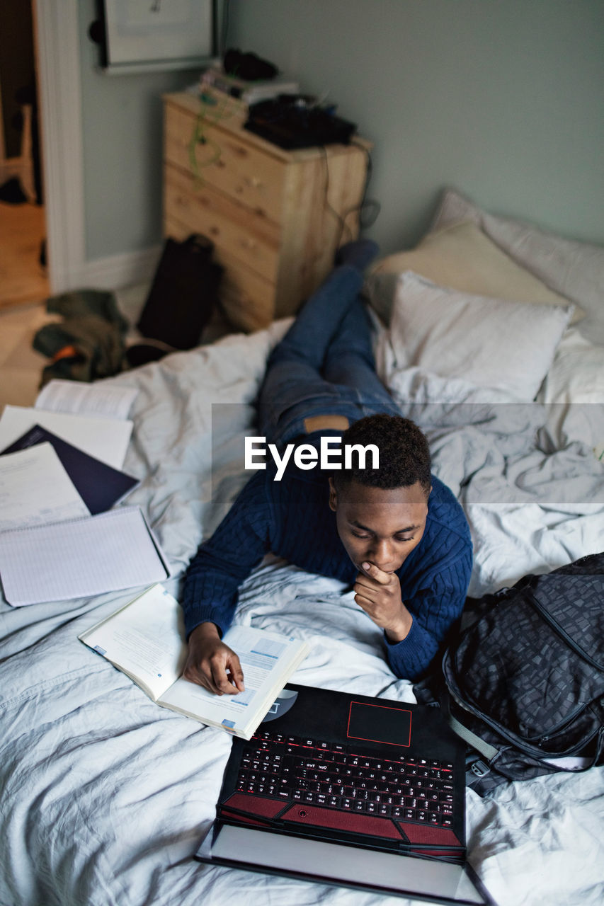 High angle view of teenage boy reading book while using laptop on bed at home