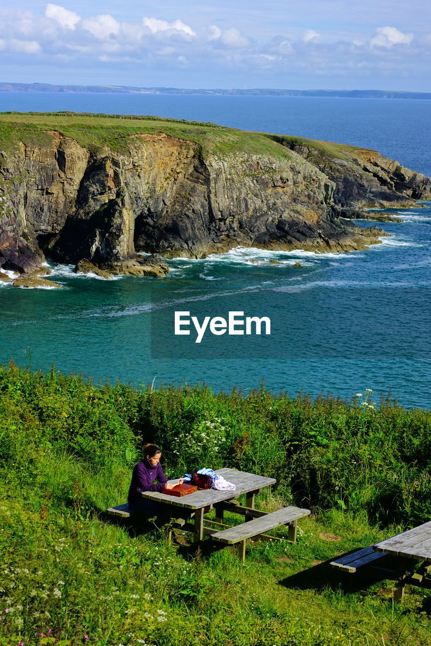 High angle view of woman sitting on picnic table by sea against cloudy sky