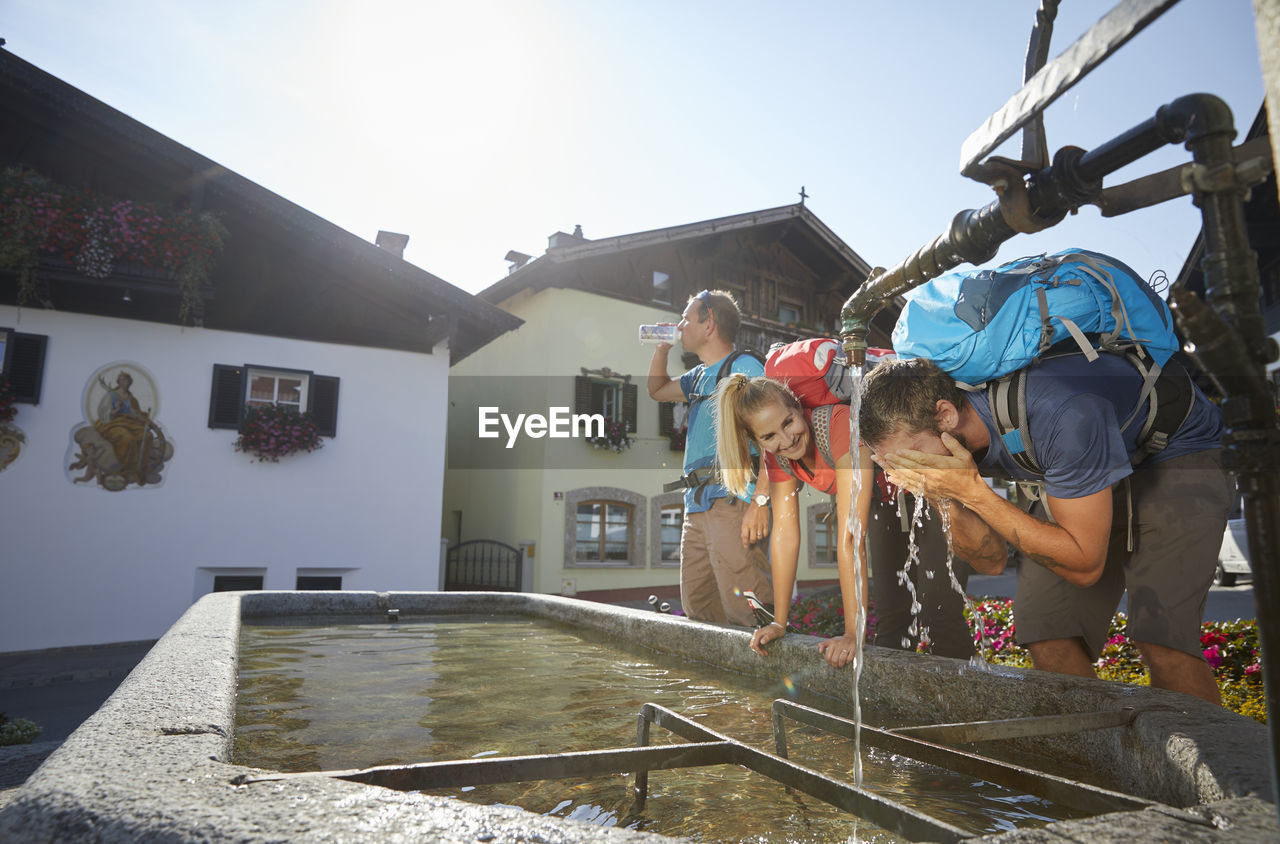 Hiker washing face by friends at fountain in village on sunny day, mutters, tyrol, austria