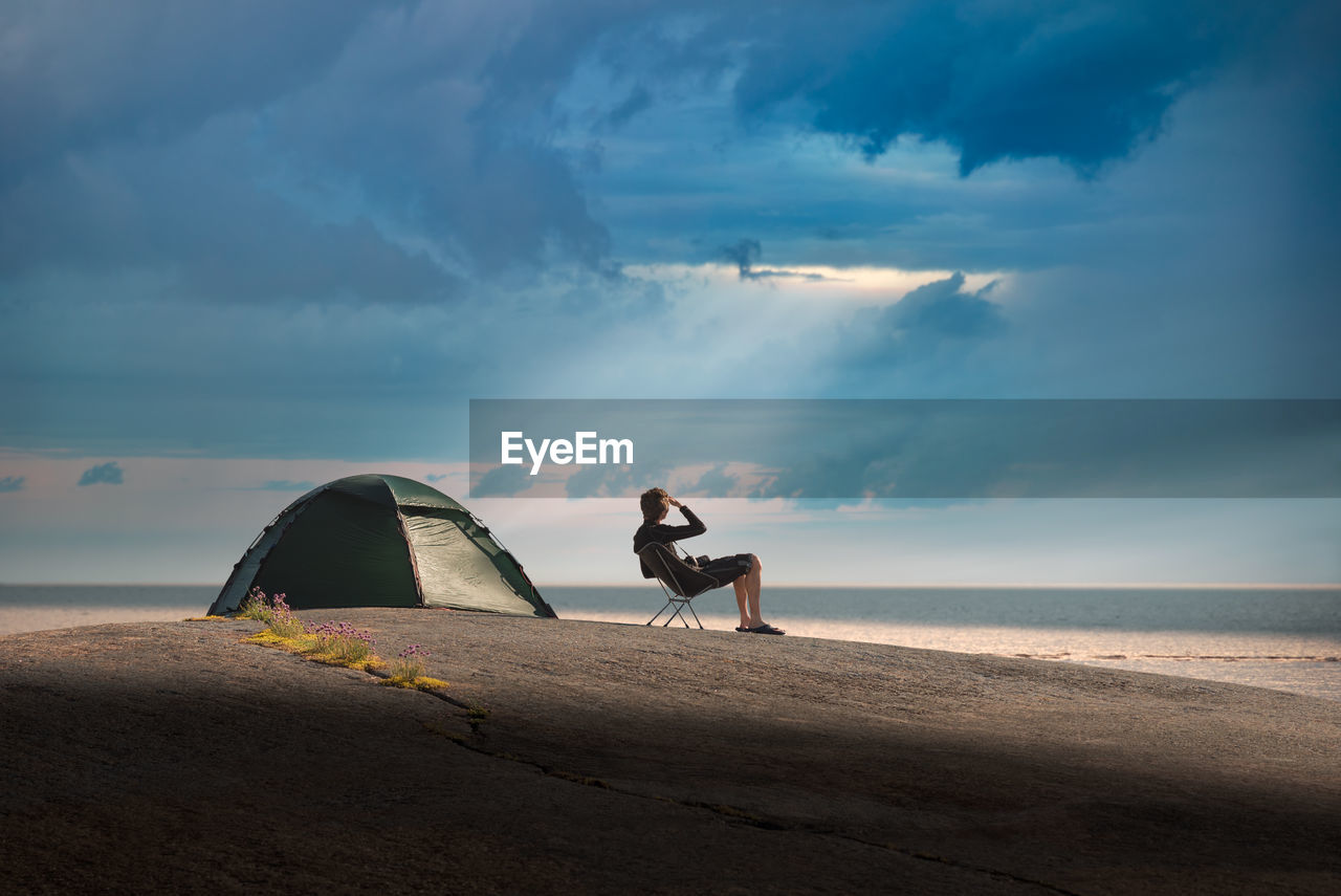 Man camping at beach against cloudy sky during sunset