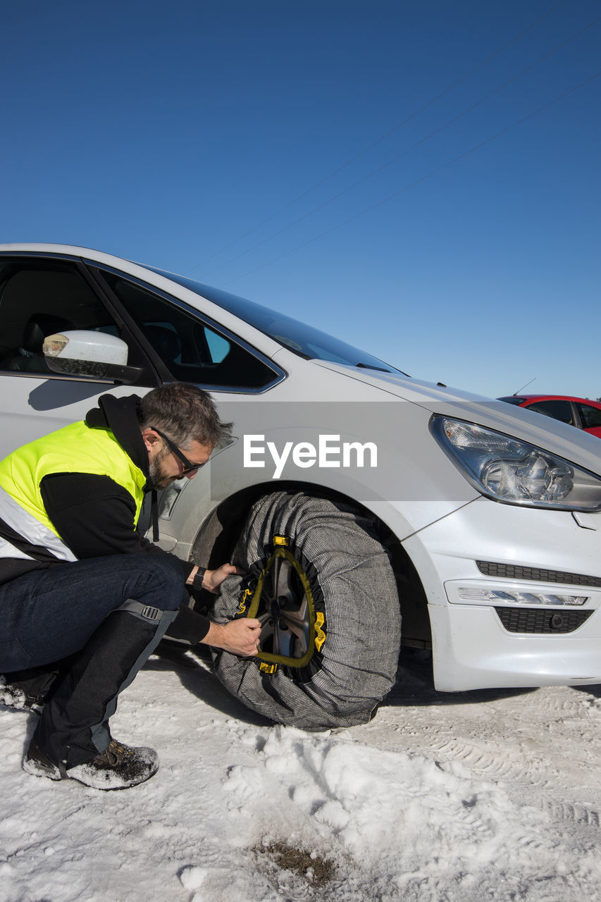 Male mechanic helping female driver with changing wheel of modern automobile on snowy road in winter