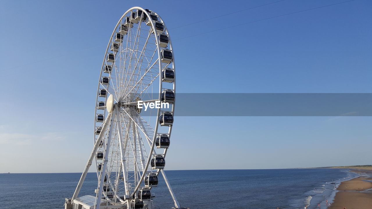 LOW ANGLE VIEW OF FERRIS WHEEL BY SEA AGAINST CLEAR BLUE SKY