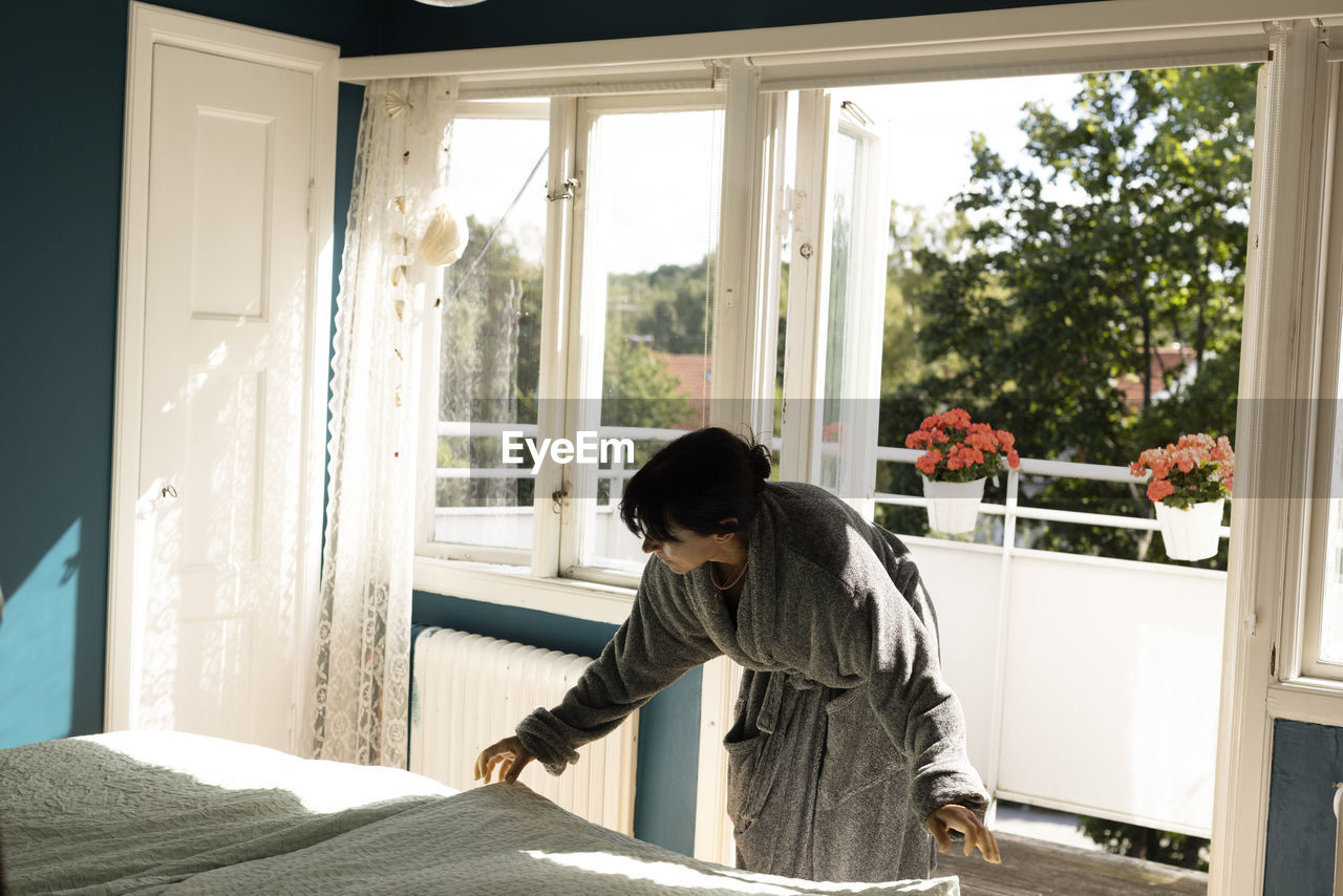 Woman wearing bathrobe laying bedsheet on bed bending against balcony doorway at home