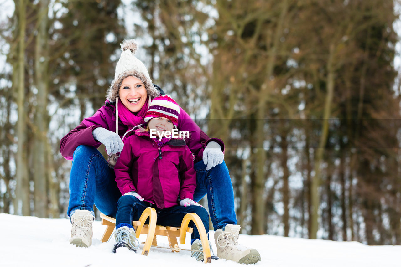 Portrait of smiling woman with daughter sitting on sled at snow covered land during winter