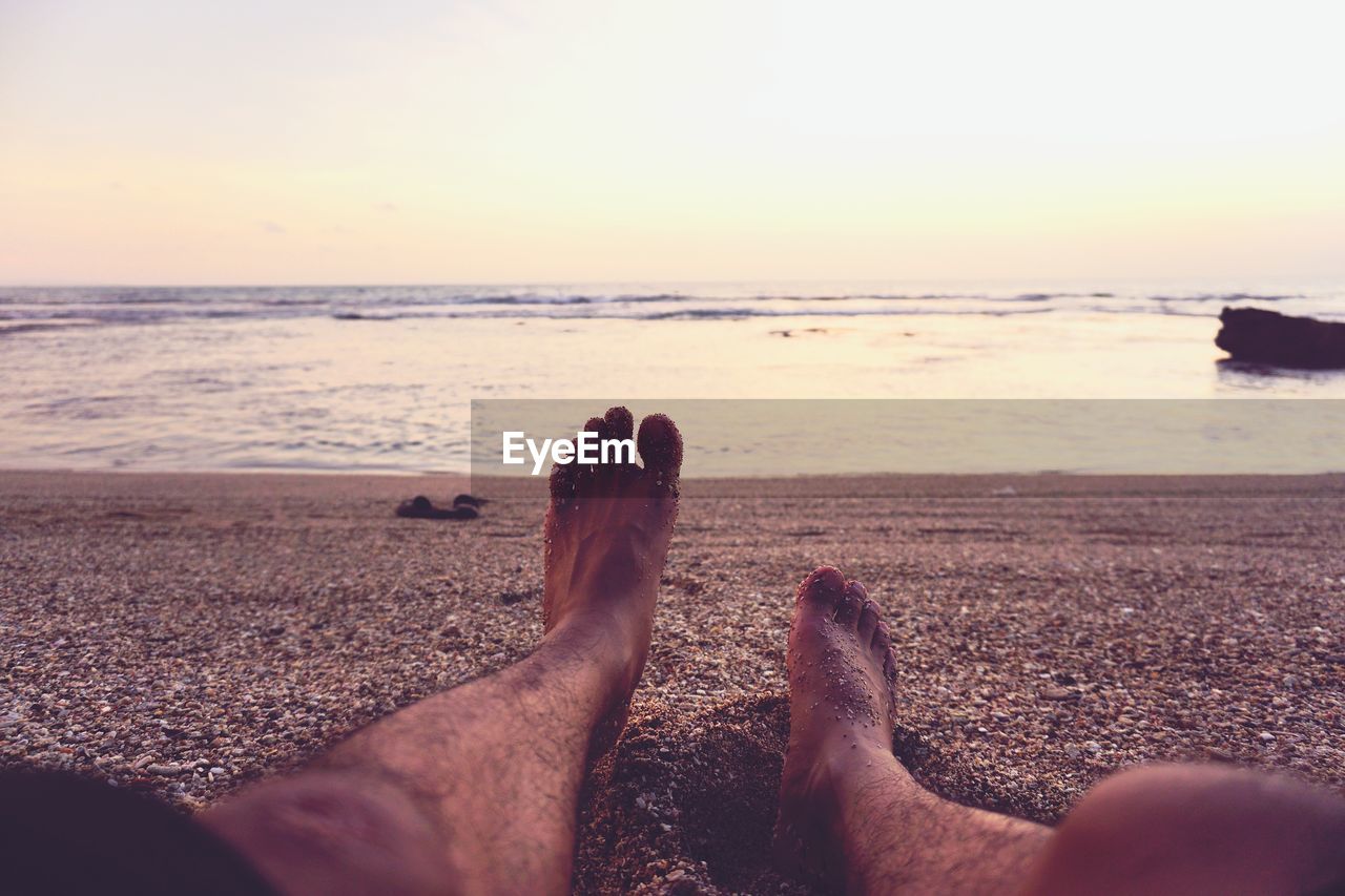 Low section of man relaxing at beach against sky during sunset