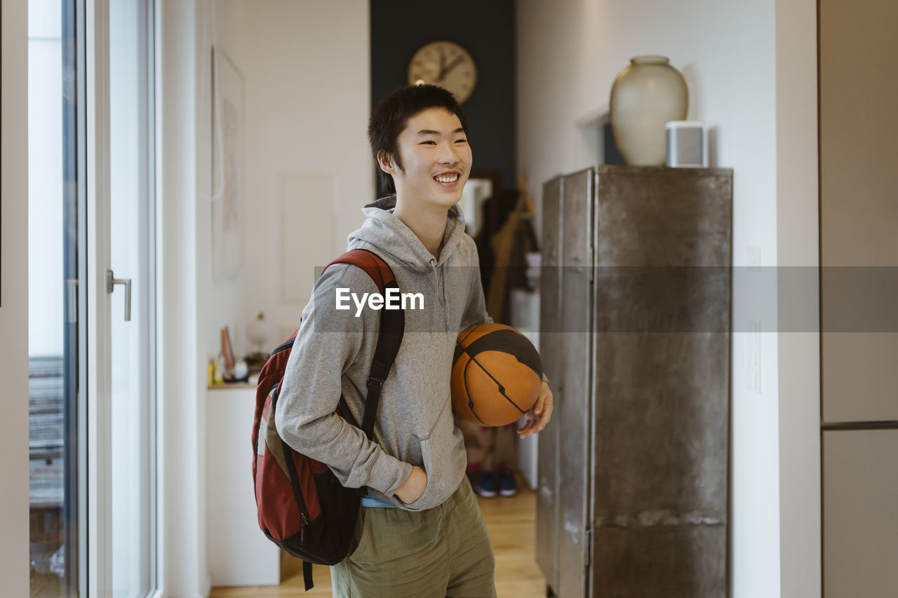 Smiling boy with hand in pocket holding basketball while standing at home