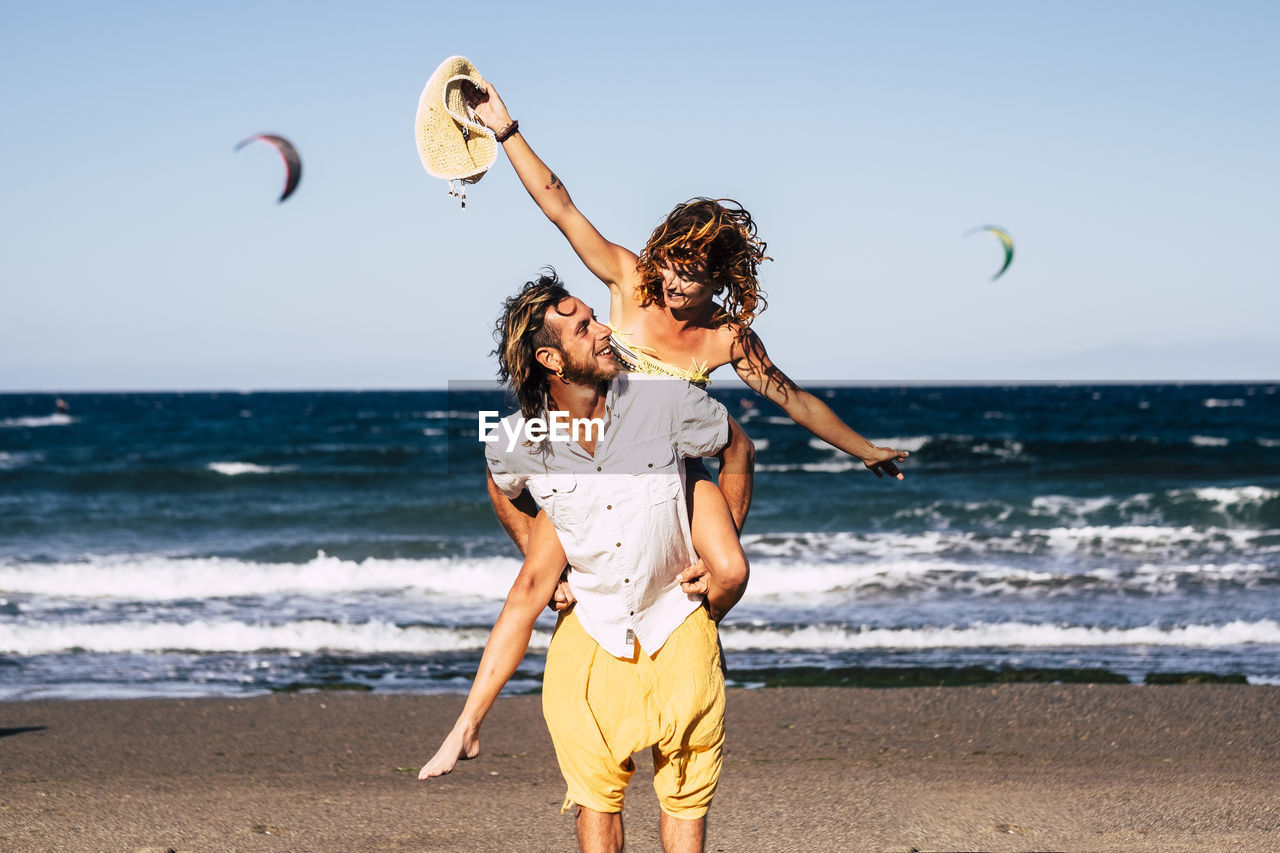 Man piggybacking cheerful woman at beach against sky