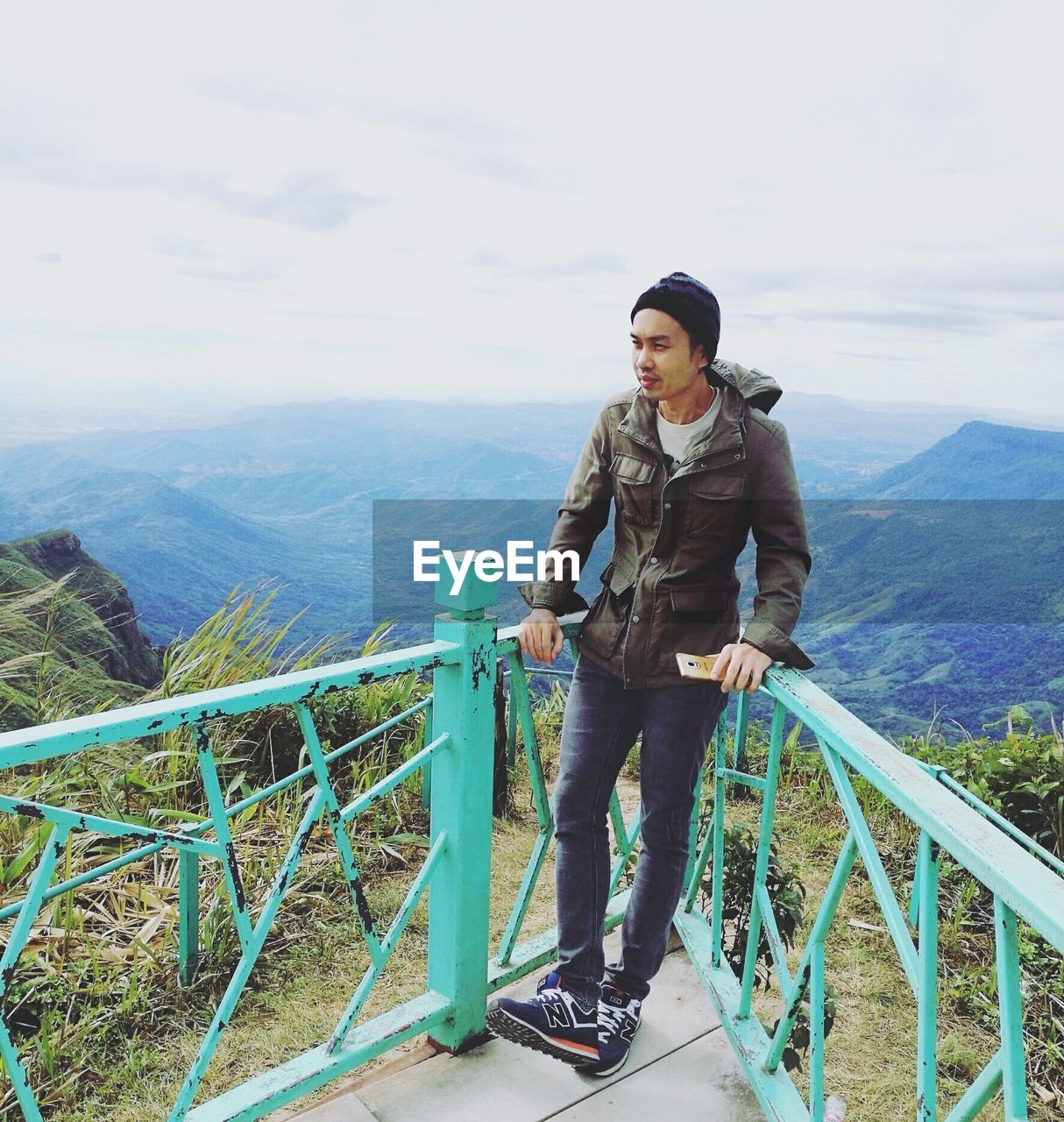 FULL LENGTH OF YOUNG WOMAN STANDING ON RAILING AGAINST MOUNTAIN