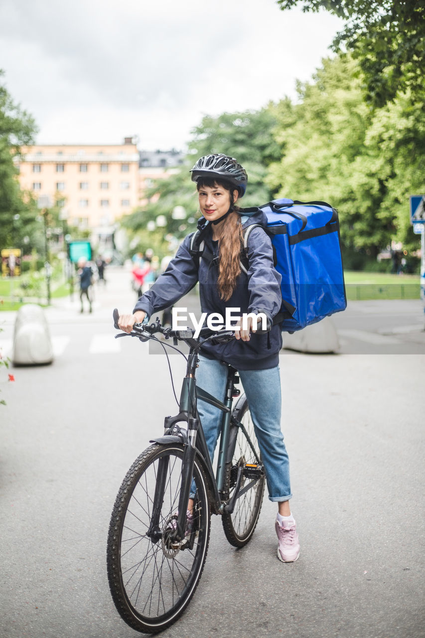 Portrait of confident food delivery woman with bicycle on street in city