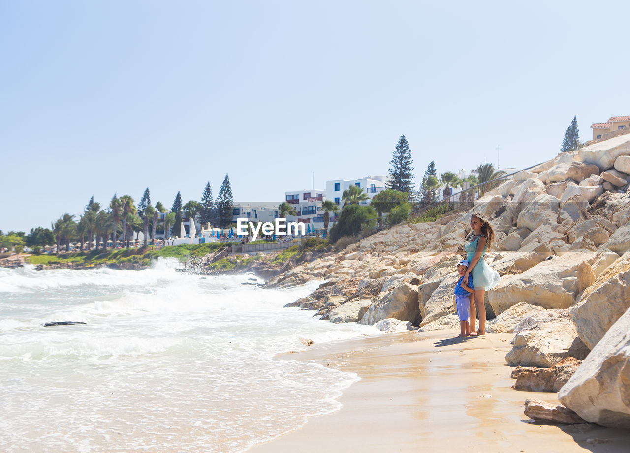 PEOPLE WALKING ON BEACH BY SEA AGAINST CLEAR SKY